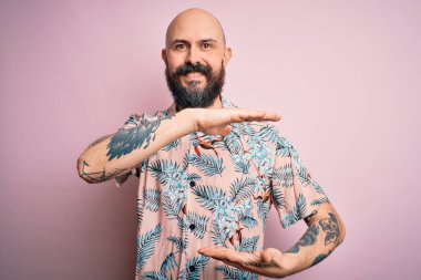 Sakallı ve dövmeli yakışıklı bir adam pembe arka planda sıradan bir çiçek gömleği giyiyor. Ellerinde büyük ve büyük beden işareti, ölçü işareti var. Kameraya bakıp gülümsüyor. Ölçüm konsepti.