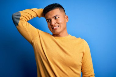 Sarı günlük süveter giyen genç, yakışıklı latin adam mavi arka planda gülümsüyor, el hareketleriyle saçlara dokunuyor, çekici ve şık poz veriyor.