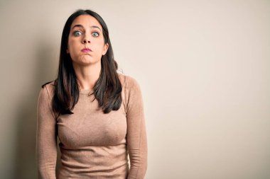 Mavi gözlü genç esmer kadın izole edilmiş beyaz arka planda komik yüzlü yanakları şişiren günlük süveter giyiyor. Havayla şişmiş ağız, çılgın bir ifade..