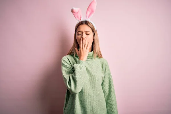 年轻美丽的女人戴着东方兔子耳朵站在孤立的粉红色背景上 厌烦了打呵欠累了用手捂住的嘴 烦躁不安和困倦 — 图库照片