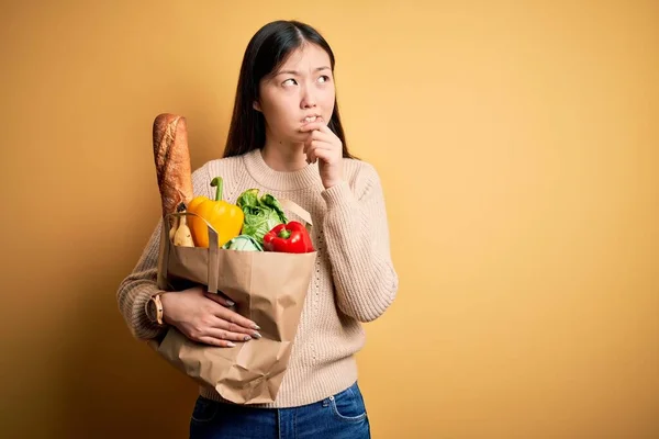 年轻的亚洲女人手里拿着一袋袋新的健康食品放在黄色孤立的背景下思考着一个问题 手托下巴忧心忡忡 — 图库照片