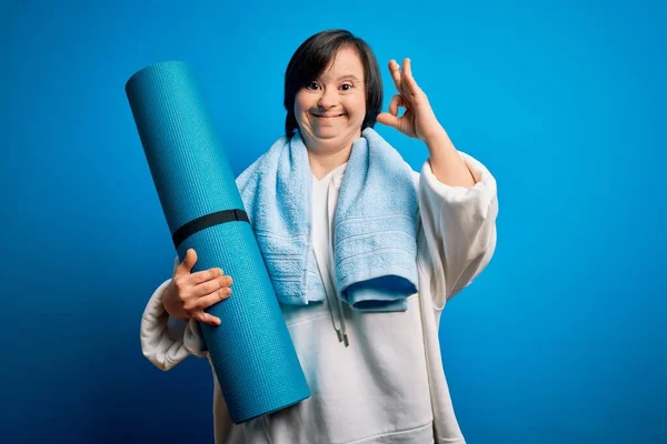 Young down syndrome fitness woman training yoga and pilates holding workout mat doing ok sign with fingers, excellent symbol