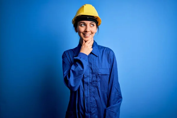 青い目をした若い美しい労働者の女性は セキュリティヘルメットを着用し 質問について考える顎の手で一様性 集中的な表現 思慮深い顔で微笑んだ 疑いの概念 — ストック写真