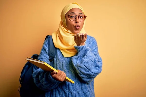 Young African American student woman wearing muslim hijab and backpack holding book looking at the camera blowing a kiss with hand on air being lovely and sexy. Love expression.