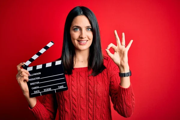Young director woman with blue eyes making movie holding clapboard over red background doing ok sign with fingers, excellent symbol