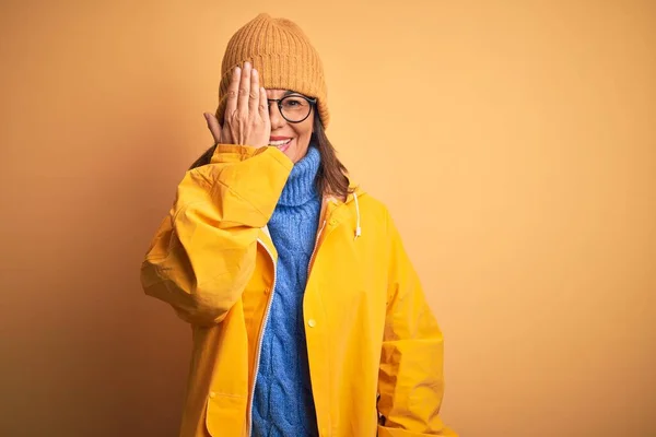 中年女性身に着けています黄色のレインコートと冬の帽子上の隔離された背景カバー1目で手 顔に自信を持って笑顔と驚きの感情 — ストック写真