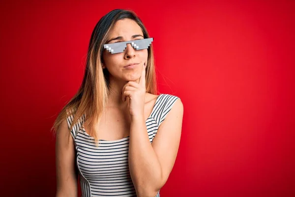 Young beautiful brunette woman wearing funny thug life sunglasses over red background with hand on chin thinking about question, pensive expression. Smiling with thoughtful face. Doubt concept.
