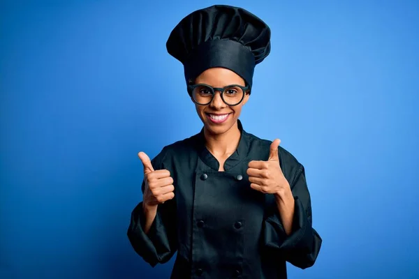 年轻的非洲女厨师身穿炊具制服 头戴蓝色背景的帽子 手握双手 竖起大拇指 微笑着 快乐地做着积极的手势 快乐的表情和胜利的姿态 — 图库照片