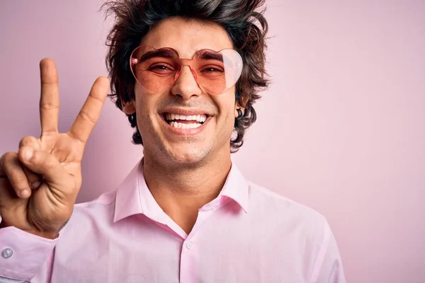 孤立したピンクの背景の上にハートの形をしたメガネをかけている若いハンサムな男が 勝利のサインをしているカメラで幸せそうな顔をして笑っている 第二番 — ストック写真