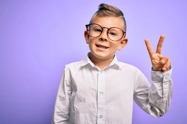 青い目をした若い白人の子供が眼鏡をかけ 紫色の背景に白いシャツを着て カメラで勝利のサインをして幸せな顔をして笑っています 第二番 — ストック写真