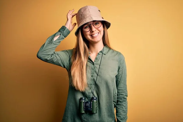 Beautiful blonde explorer woman with blue eyes wearing hat and glasses using binoculars confuse and wonder about question. Uncertain with doubt, thinking with hand on head. Pensive concept.