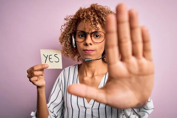 African american call center agent woman using headset holding reminder with yes message with open hand doing stop sign with serious and confident expression, defense gesture