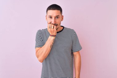 Pembe izole edilmiş arka plan üzerinde duran çizgili tişört giyen yakışıklı genç adam elleri ağız yiyen tırnaklarla gergin ve stresli görünüyor. Endişe sorunu.