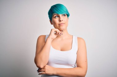 Mavi saçlı, beyaz arka planda sıradan tişörtler giyen, elleri çenesinde soru soran, düşünceli bir kadın. Gülen ve düşünceli bir yüz. Şüphe kavramı.