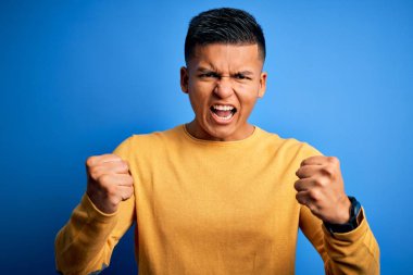 İzole edilmiş mavi arka plan üzerine sarı süveter giyen genç ve yakışıklı Latin adam öfkeyle bağırırken kızgın ve öfkeli yumruklar sallıyor. Öfke ve agresif kavram.