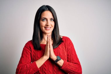 Young brunette woman with blue eyes wearing casual sweater over isolated white background praying with hands together asking for forgiveness smiling confident.