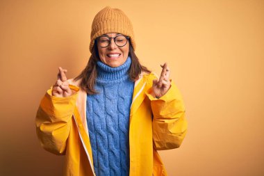 Sarı yağmurluk ve kış şapkası takan orta yaşlı bir kadın izole edilmiş arka planda umut ve gözler kapalı gülümseyerek el sallıyor. Şans ve batıl inanç..