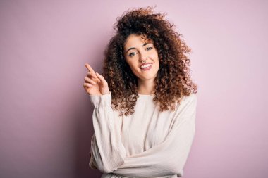 Kıvırcık saçlı ve piercingli güzel bir kadın pembe arka planda rahat bir süveter giyiyor ve bir numaralı parmağını gösterip gülümserken mutlu ve kendinden emin..