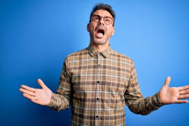 Mavi arka planda gözlük takan genç, yakışıklı bir adam. Deli deli bağırıyor, bağırıyor, agresif bir ifade ve kollarla bağırıyor. Hayal kırıklığı konsepti.