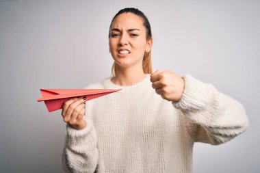 Mavi gözlü güzel sarışın kadın beyaz arka planda kırmızı kağıt uçak tutuyor sinirlenmiş ve hayal kırıklığına uğramış öfkeyle bağırıyor, çıldırmış ve el kaldırarak bağırıyor.