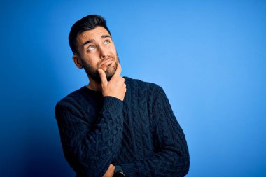 İzole edilmiş mavi arka plan üzerinde duran sıradan bir süveter giyen genç yakışıklı bir soru hakkında endişeli ve gergin bir şekilde düşünüyor.