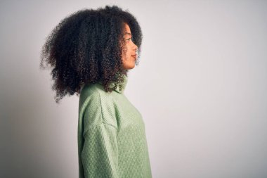 Afro saçlı, yeşil kışlık kazak giyen genç ve güzel Afro-Amerikalı kadın. Yan tarafa bakıyor, rahat poz veriyor, kendine güvenen bir gülümsemesi var..
