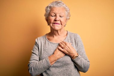 Üst düzey güzel bir kadın, izole edilmiş sarı arka plan üzerinde sıradan bir tişört giyip elleri göğsünde, gözleri kapalı ve yüzünde minnet dolu bir jest ile dikiliyor. Sağlık kavramı.