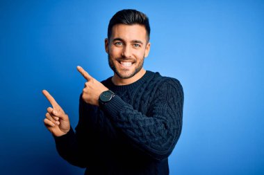 Genç, yakışıklı bir adam, sıradan bir süveter giymiş, mavi arka planda dikiliyor gülümsüyor ve kameraya bakıyor, iki eliyle ve parmaklarıyla yan tarafa bakıyor..