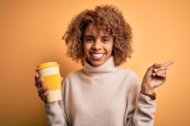 Genç Afro-Amerikan kıvırcık kadın sarı arka planda bir fincan kahve içiyor.