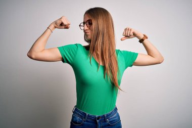 Genç, güzel, kızıl saçlı, sıradan yeşil tişörtler giyen ve beyaz arka planda gözlük takan bir kadın gururla gülümseyen kol kaslarını gösteriyor. Fitness konsepti.