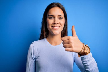 Young beautiful brunette woman wearing casual sweater standing over blue background doing happy thumbs up gesture with hand. Approving expression looking at the camera showing success.