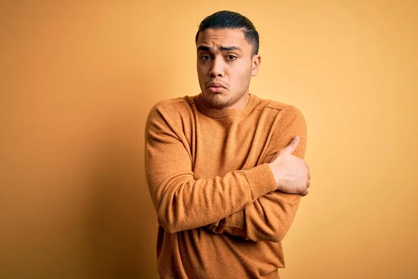 Young brazilian man wearing casual sweater standing over isolated yellow background shaking and freezing for winter cold with sad and shock expression on face