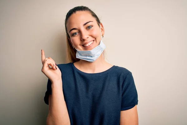 Beautiful blonde woman with blue eyes wearing medical mask over white background with a big smile on face, pointing with hand and finger to the side looking at the camera.