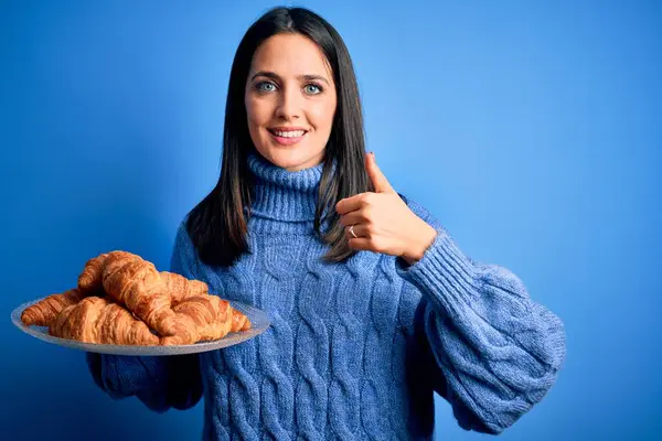 年轻的女人 蓝眼睛 带着甜羊角面包在孤独的背景下吃早餐 开心的笑着做个好手势 用手指竖起大拇指 很好的标志 — 图库照片