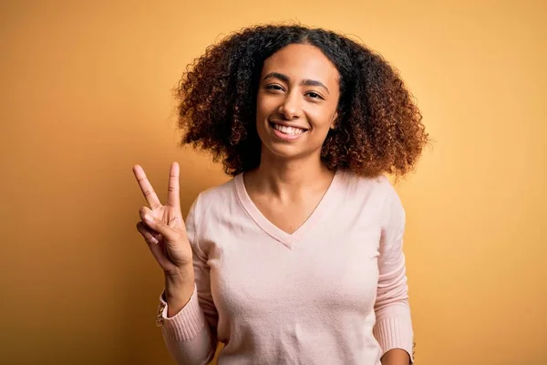 黄色の背景の上にカジュアルなセーターを着てアフロの髪を持つ若いアフリカ系アメリカ人女性は 勝利のサインを行うカメラで幸せな顔のウィンクで笑顔 第二番 — ストック写真