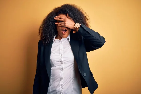 Young beautiful african american business woman with afro hair wearing elegant jacket peeking in shock covering face and eyes with hand, looking through fingers with embarrassed expression.