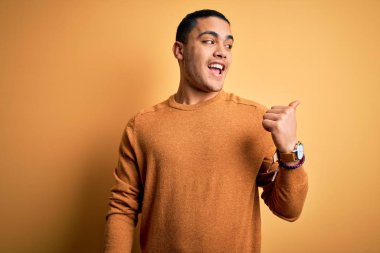 İzole edilmiş sarı arka planda dikilen sıradan bir süveter giyen genç Brezilyalı adam gülümsüyor ve başparmağıyla yan tarafa bakıyor..