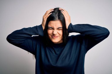 Genç, güzel, esmer kadın beyaz arka planda dikilip acı ve migren yüzünden baş ağrısı çeken sıradan bir kazak giyiyor. Ellerini başının üstüne koy..