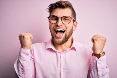 Sakallı, mavi gözlü, pembe tişörtlü ve gözlüklü genç, yakışıklı sarışın adam kolları havada ve açık bir şekilde başarıyı kutluyor ve şaşırıyor. Kazanan konsept.