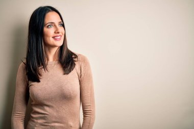 Young brunette woman with blue eyes wearing casual sweater over isolated white background looking away to side with smile on face, natural expression. Laughing confident.