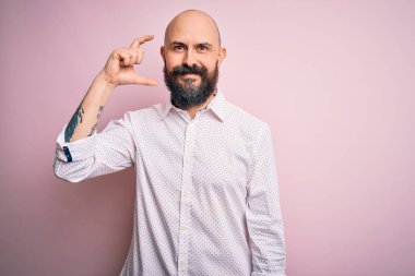 Sakallı, sakallı, zarif tişörtlü, soyutlanmış pembe arka planda gülümseyen, kendine güvenen, el hareketleriyle küçük boy işareti yapan, parmaklarıyla işaret eden ve kameraya bakan yakışıklı bir adam. Ölçü kavramı.
