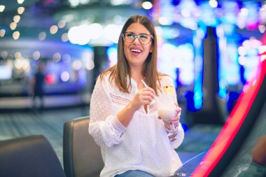 Tatilde mutlu ve kendinden emin gülümseyen genç ve güzel bir kadın. Yüzünde gülümsemeyle oturmuş kokteyl içiyor ve kumarhanede kumar oynuyor.