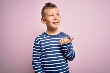 Mavi gözlü, pembe arka planda deniz çizgili tişört giyen mutlu yüzüyle gülümseyen ve baş parmağıyla yan tarafı gösteren küçük beyaz bir çocuk..