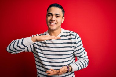 Sıradan çizgili tişört giyen genç Brezilyalı adam izole edilmiş kırmızı arka planda duruyor. Ellerinde büyük ve büyük boy işareti, ölçü işareti var. Kameraya bakıp gülümsüyor. Ölçüm konsepti.