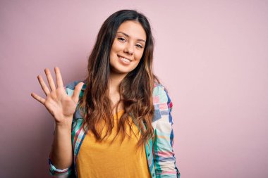 Genç, güzel, esmer, sıradan bir gömlek giyen, pembe arka planda duran, beş numarayı gösteren, kendinden emin ve mutlu gülümseyen bir kadın..