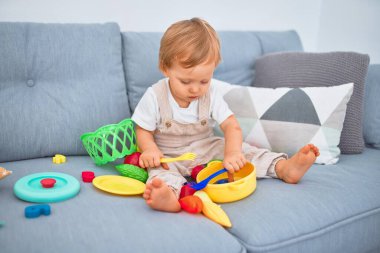Sevimli sarışın çocuk kanepede oturup evde plastik yemek oyuncaklarıyla oynuyor.