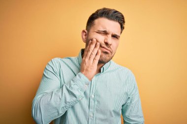 Sarı arka plan üzerine zarif yeşil gömlek giyen genç bir iş adamı diş ağrısı ya da diş hastalığından dolayı acı dolu bir ifadeyle ağıza dokunuyor. Dişçi