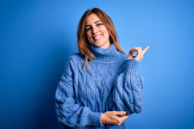 Mavi arka planda balıkçı yaka kazak giyen genç, güzel, esmer bir kadın. Yüzünde kocaman bir gülümseme, eli ve parmağı kameraya bakıyor..