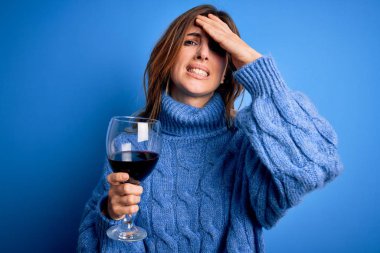 Genç, güzel esmer kadın, izole edilmiş mavi arka planda bir bardak kırmızı şarap içerken, elleri başının üstünde, utanç ve şaşkınlık dolu, sinirli ve hayal kırıklığına uğramış bir halde. Hata için korku ve üzüntü.