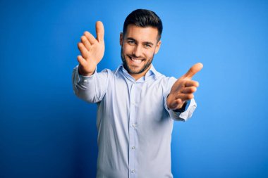 Zarif gömlekli genç adam, izole edilmiş mavi arka planda dikilip kameraya bakarak sarılmak için kollarını açmış gülümsüyor. Mutluluğu kucaklayan neşeli bir ifade..